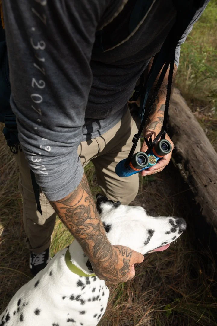 A person with tattoos on their arm holds a Dalmatian dog by its collar while exploring a forest with a hand-held binoculars hanging around their neck.