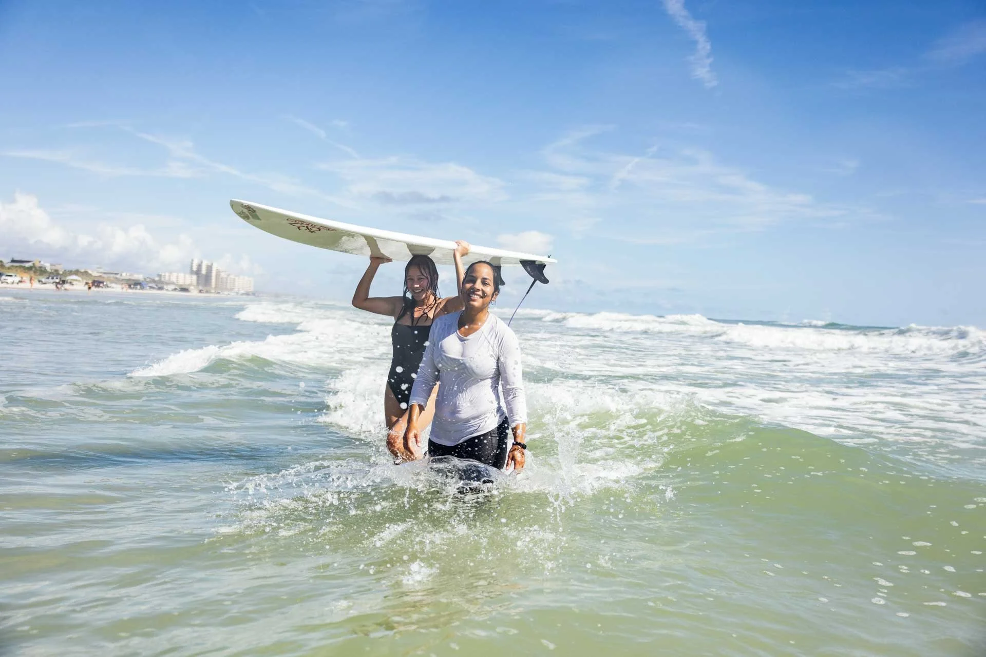 Two women in the ocean, one holding a surfboard, enjoying a sunny day at the beach.
