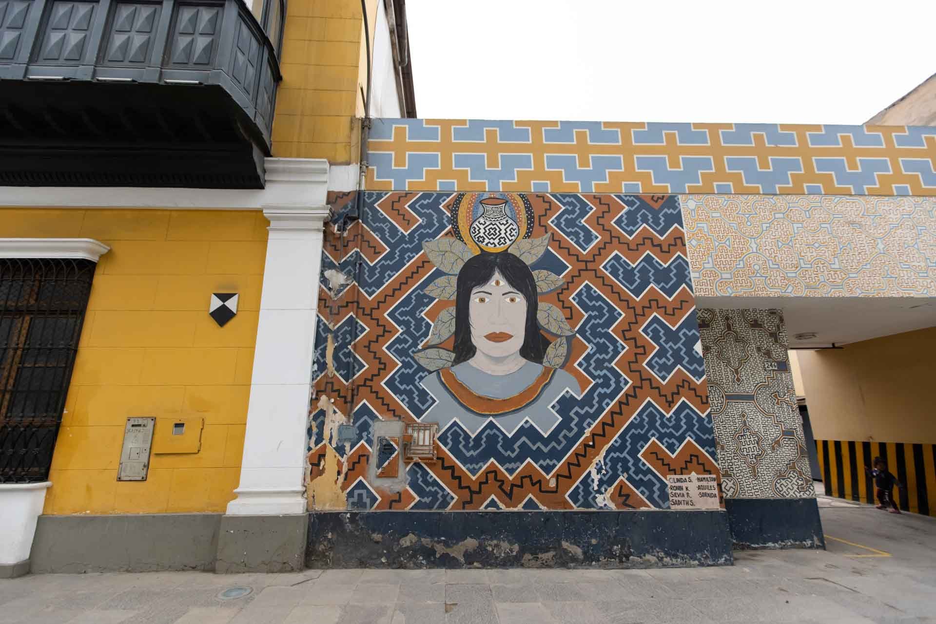 Colorful mural on an exterior building wall depicting a woman with dark hair, wearing a necklace, surrounded by geometric patterns and a jar on top of her head, with a background of various intricate designs and patterns.