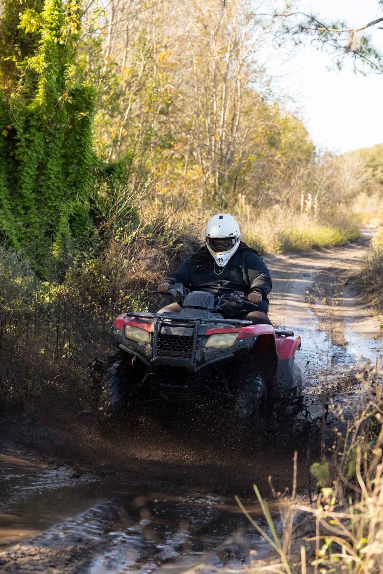 Person riding an ATV through a muddy trail surrounded by trees and bushes.