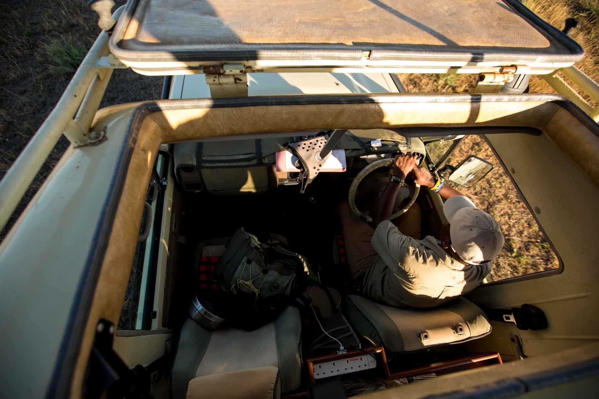 Overhead view of a person driving an off-road vehicle through a dirt area during daylight, wearing a cap and wristbands, with backpacks and equipment inside the vehicle.