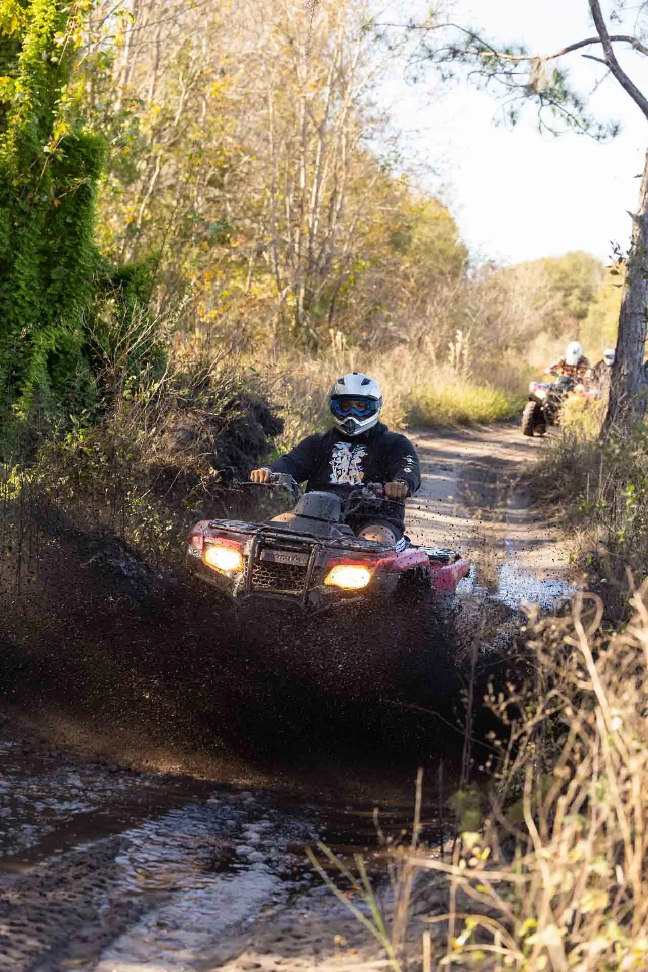A person riding an ATV through muddy terrain in a wooded outdoor area, with two more ATV riders visible in the background.