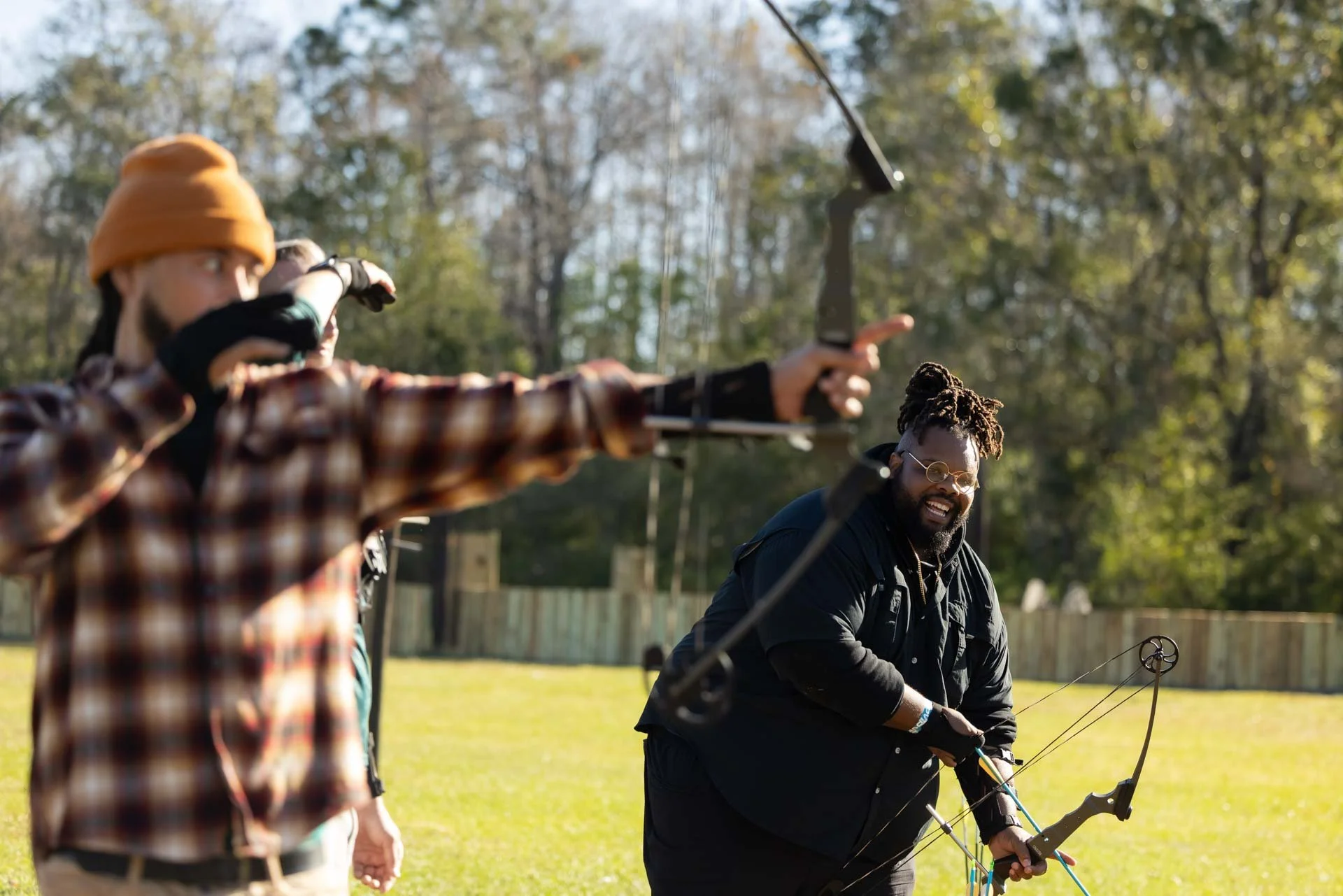 A group of people outdoors, with one person aiming a bow and arrow while others watch, surrounded by trees and a grassy field.