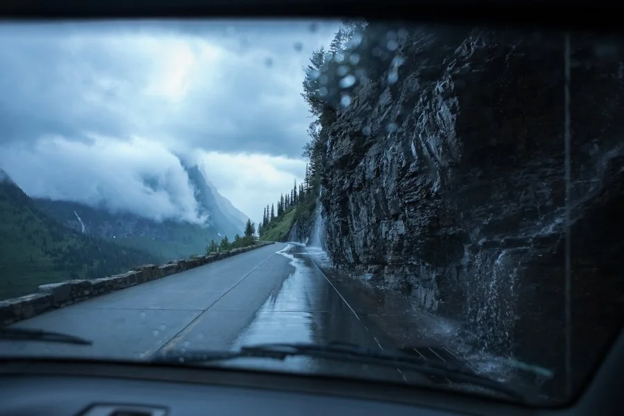 Road with rain on windshield, steep rocky cliff on the right, mountain landscape with clouds in the distance, and trees on the hillside