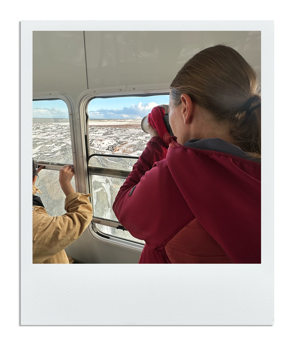 Two people on a snow-covered landscape, looking out of a window, with one using binoculars.