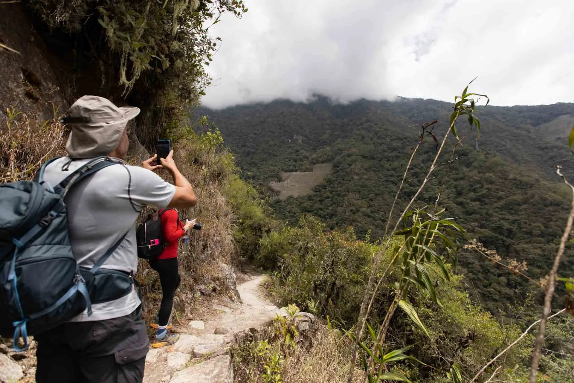 People hiking on a mountain trail, capturing photos of the lush, green mountains and cloudy sky.