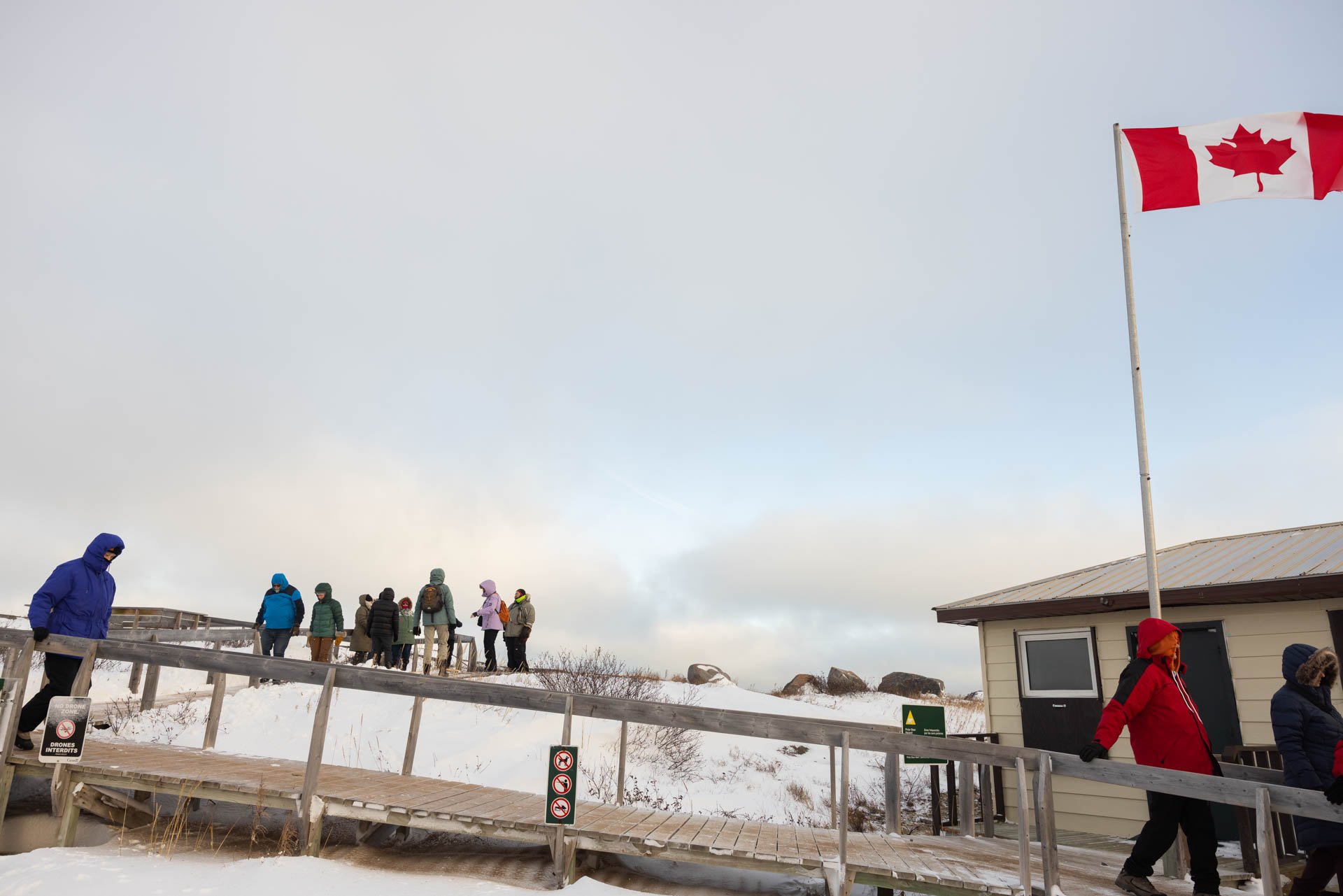 Group of people in winter clothing walking up a snowy ramp outside near a small building, with a Canadian flag flying on a flagpole.