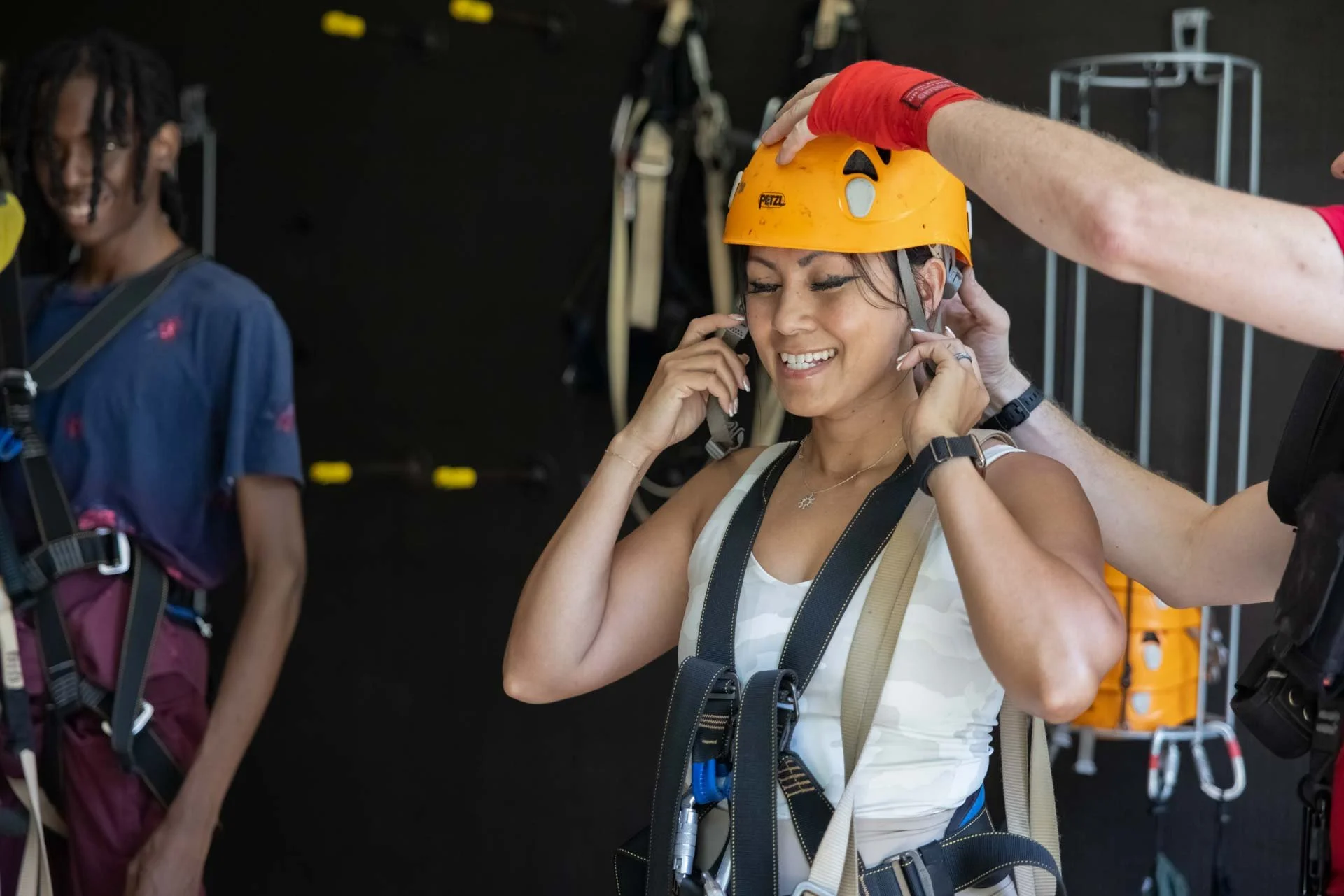 A smiling woman trying on a yellow climbing helmet while receiving assistance in a climbing gym. She is adjusting her helmet with her eyes closed, wearing a harness, and is surrounded by climbing gear.