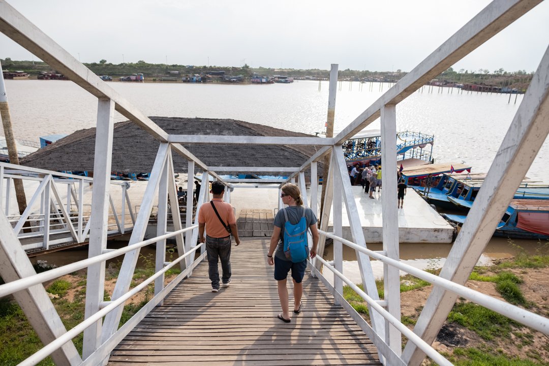 Two people walking down a wooden ramp toward a dock with boats on a lake.