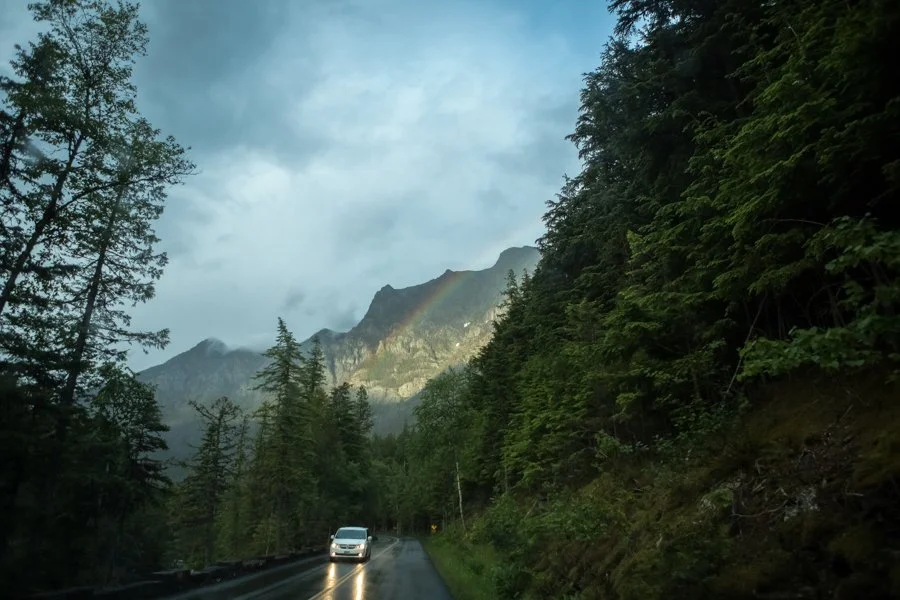 A car driving on a mountain road surrounded by tall evergreen trees with mountains and a faint rainbow in the cloudy sky.