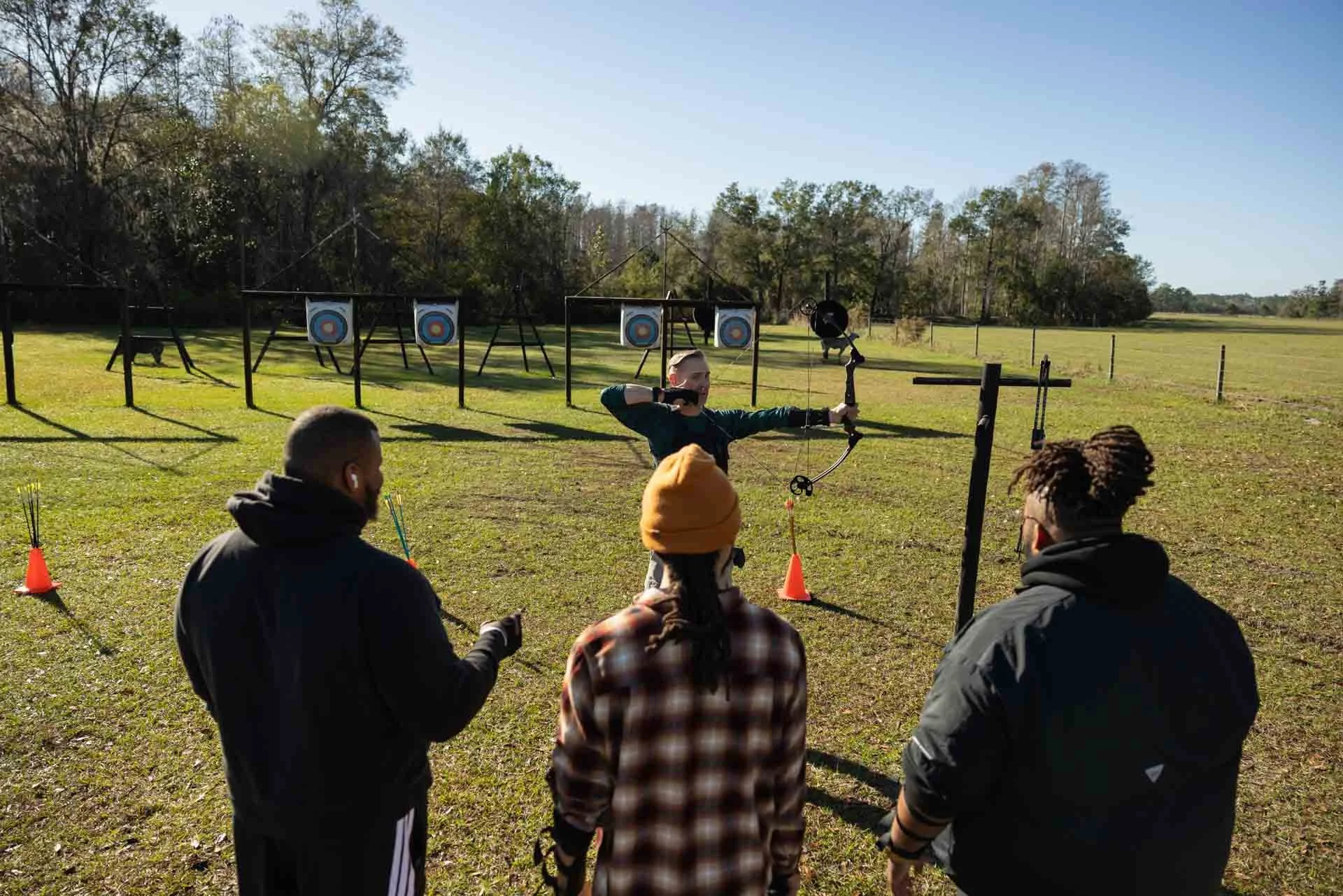 A person practicing archery outdoors with three onlookers observing, target boards in the background, and a grassy field under a clear sky.