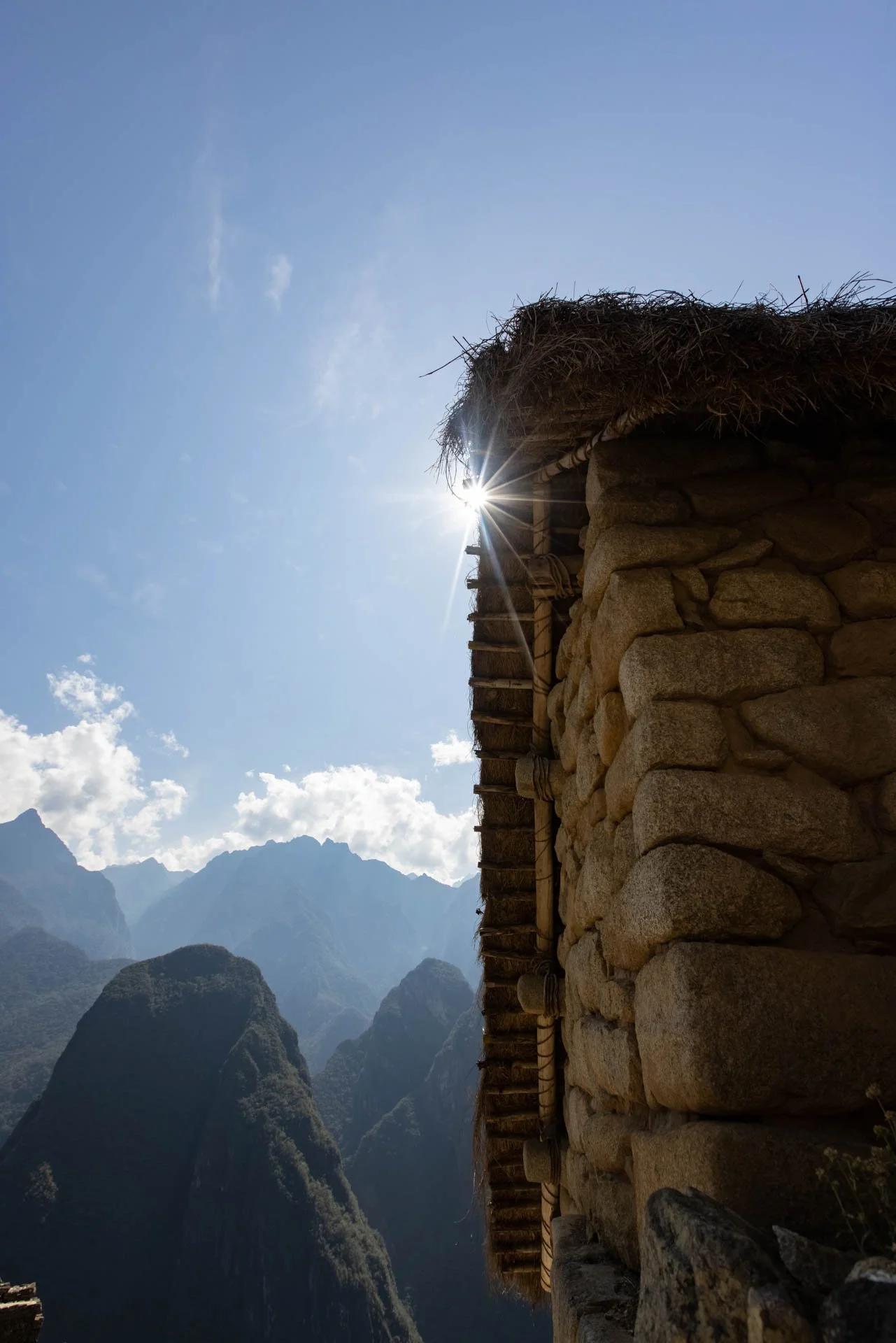 Stone building with thatched roof and mountain landscape in the background under a blue sky with the sun peeking from behind the roof.