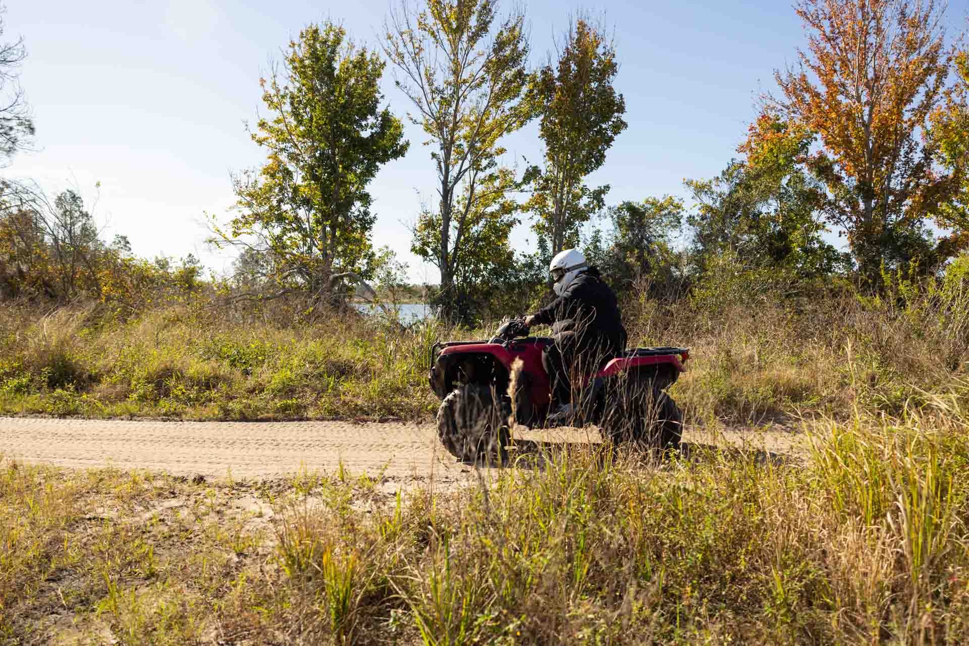 A person riding an ATV along a dirt trail in a grassy, wooded area under a clear sky.