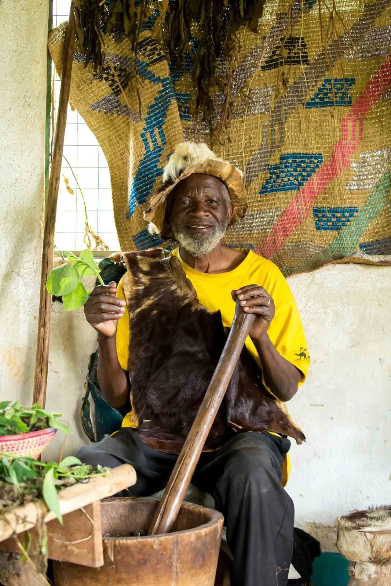 An elderly man wearing a yellow shirt, sitting indoors with a smile on his face, holding a goat. He has a head covering made of straw and a beard. The background features a colorful woven mat hanging on the wall, and the man is next to a container wi