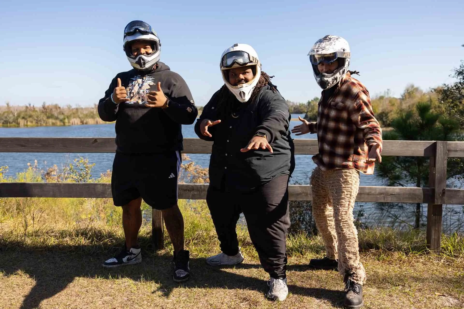 Three people in motorcycle helmets and casual clothing posing outdoors near a lake, with a wooden fence behind them and trees in the background.
