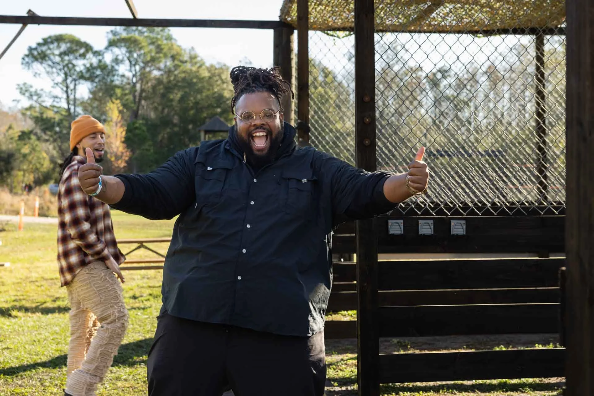 Two men standing outside near a fence, one in the foreground with arms outstretched and thumbs up, smiling widely, and the second man in the background, wearing a beanie and plaid shirt, also smiling.