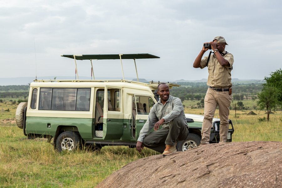 Two men with safari gear and binoculars in an open field with a green and cream-colored safari vehicle behind them.
