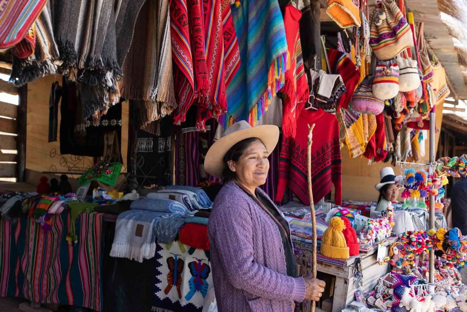 Woman with a hat selling colorful traditional textiles and souvenirs at a market stall.