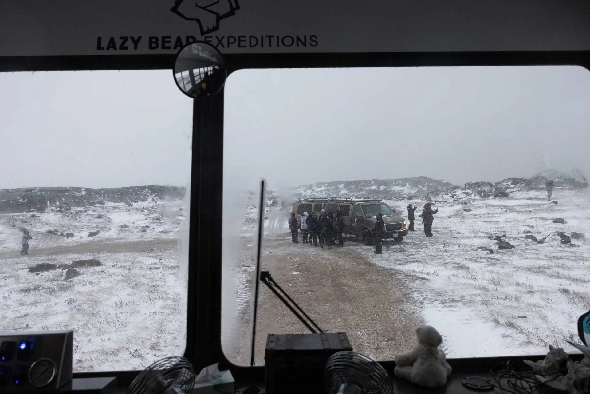 View from inside a vehicle showing a snowy landscape with a group of people and a vehicle on a dirt road outside.