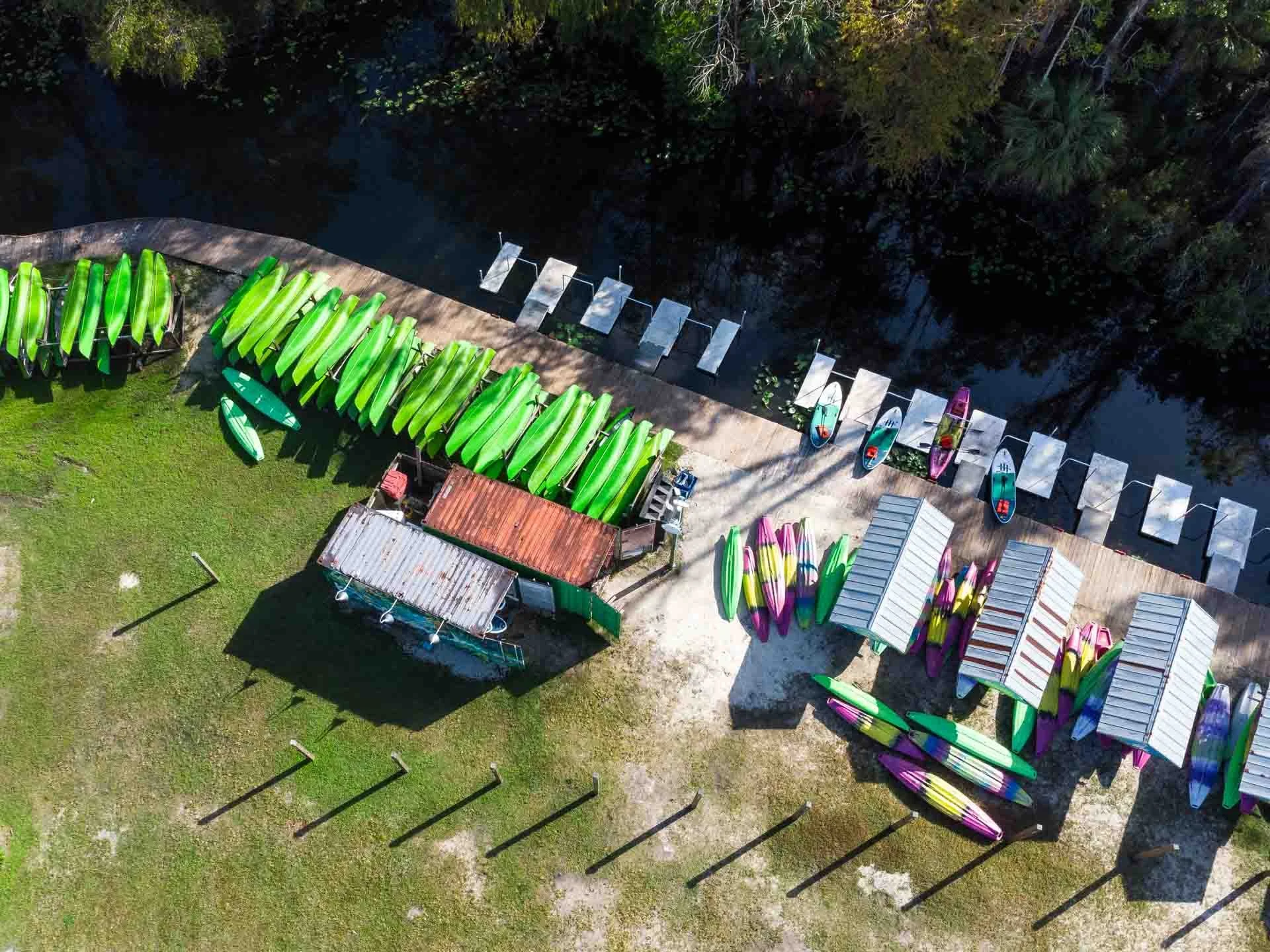An aerial view of a shoreline with colorful kayaks and paddleboards, a small dock, a building with a rusted roof, and a grassy area with shadowed pool ladders.