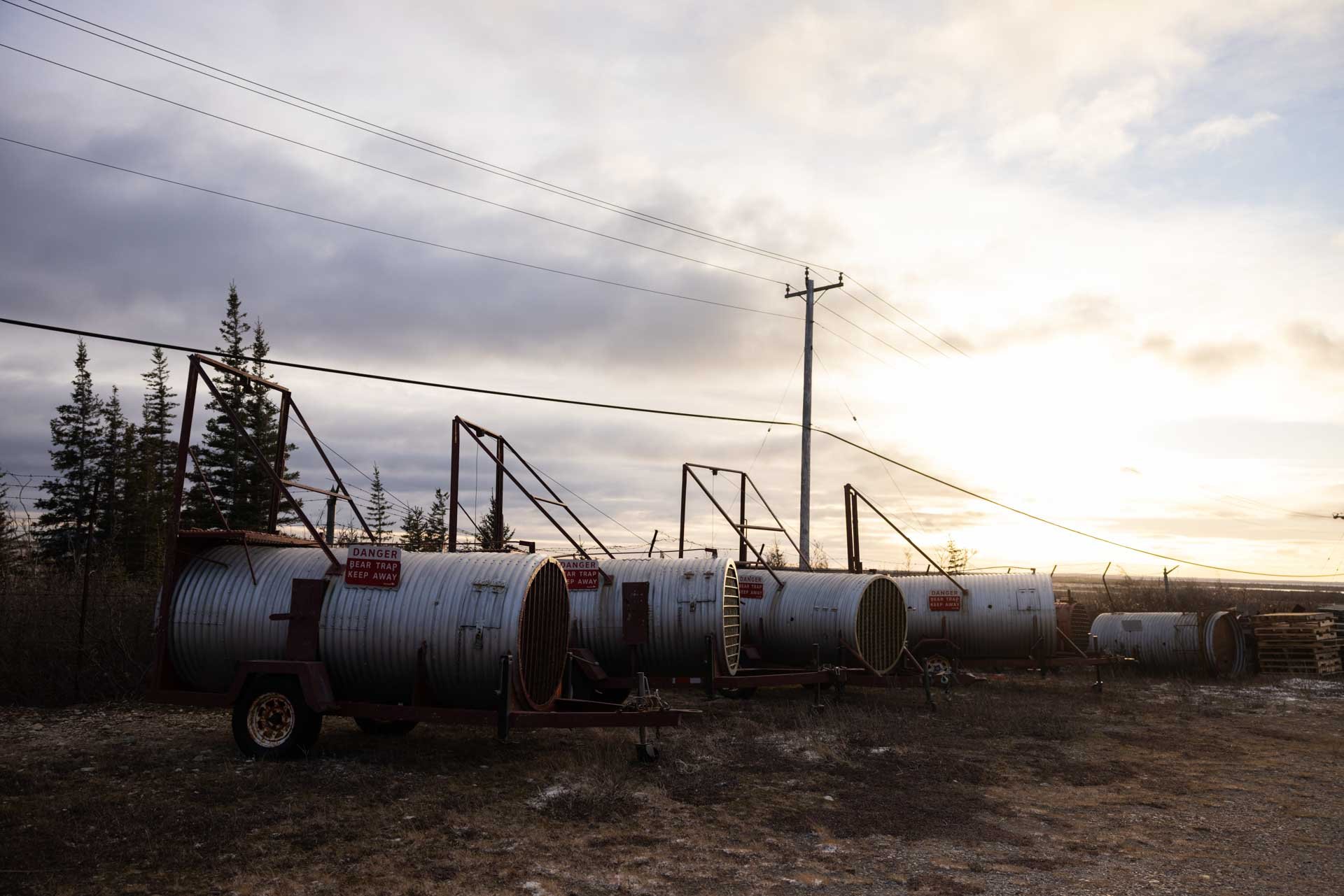 Several large metal tanks with warning signs are positioned outdoors on a dirt lot, with trees and power lines in the background. The sky is overcast with the sun low on the horizon.