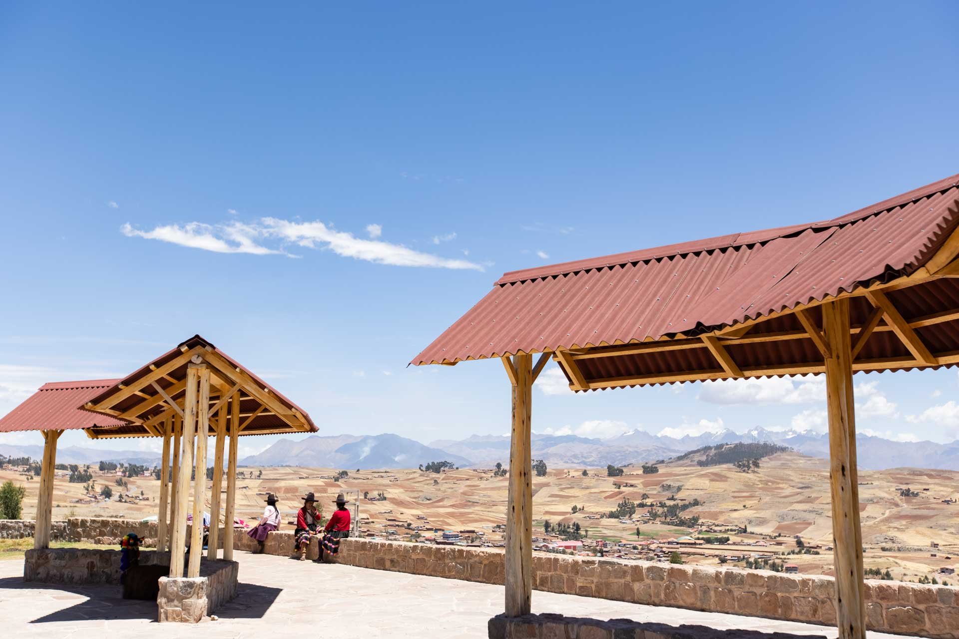 A scenic overlook with two wooden shaded structures, people sitting on a stone wall, and a vast landscape of fields and mountains under a clear blue sky.