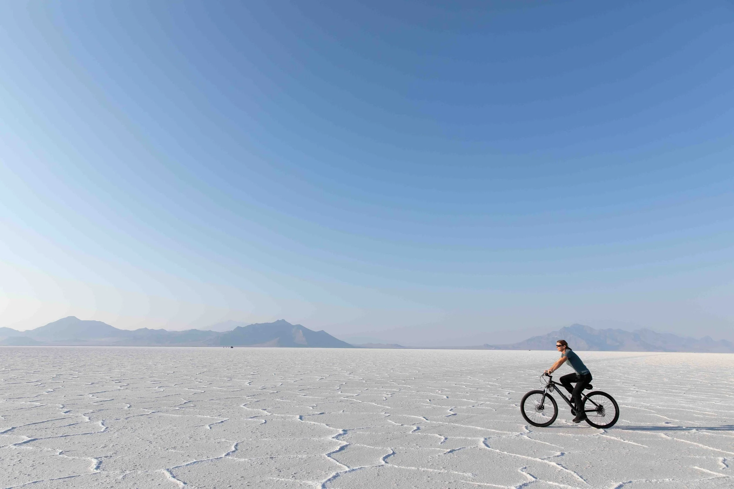 A person riding a bicycle across a vast, flat salt flat under a clear blue sky, with mountains visible in the distance.
