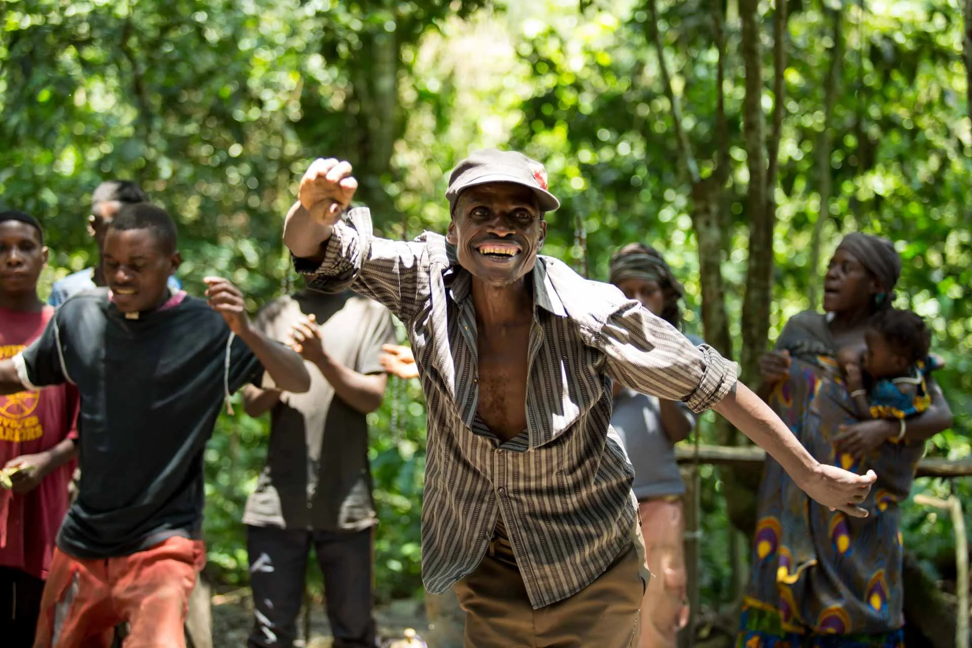 A group of people dancing and smiling in a lush green forest, with one man in the center wearing a striped shirt and a cap.