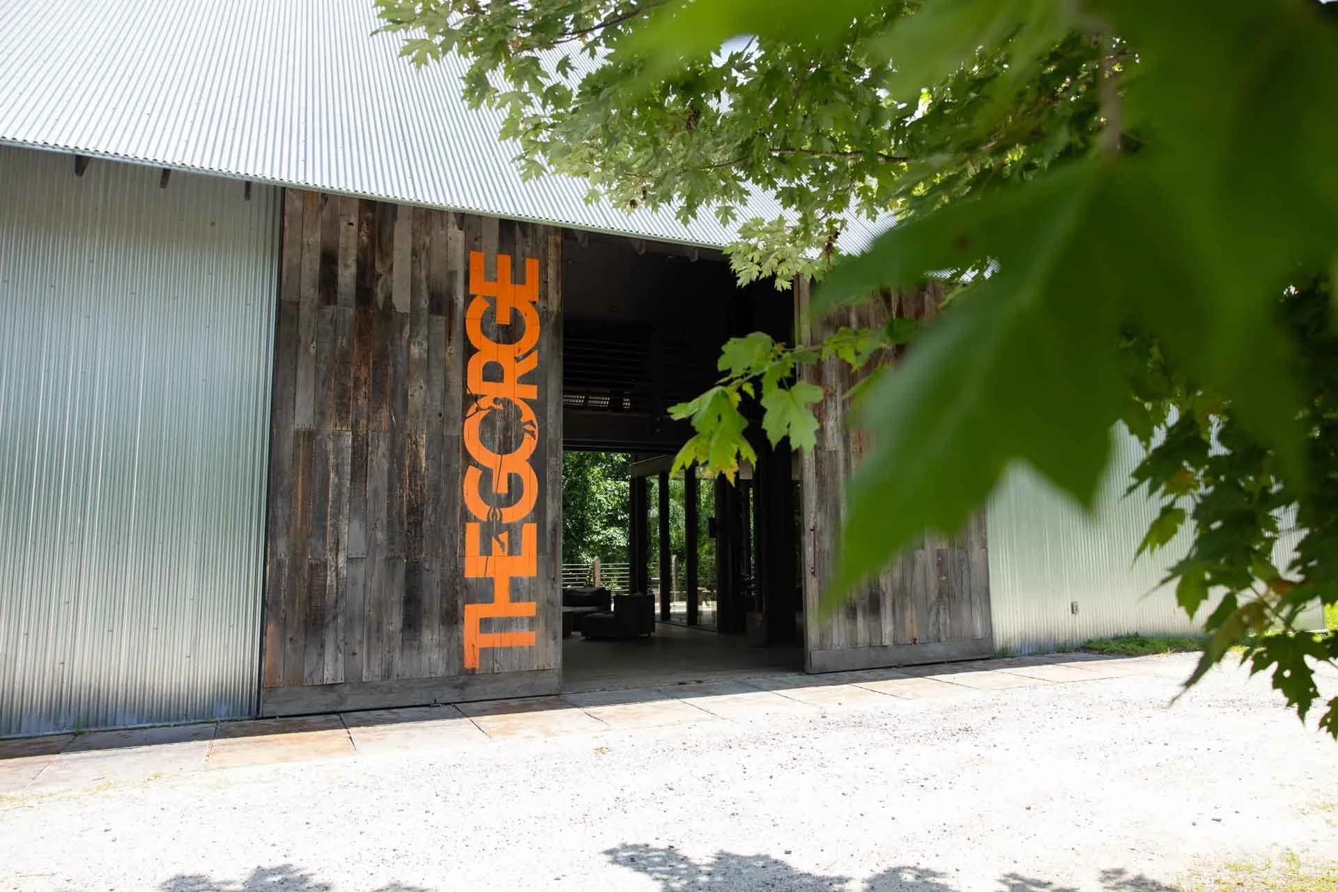 A rustic barn with a modern metal roof, wooden sliding doors, and orange lettering spelling 'THE ESCAPE' on a weathered wooden wall, surrounded by greenery.