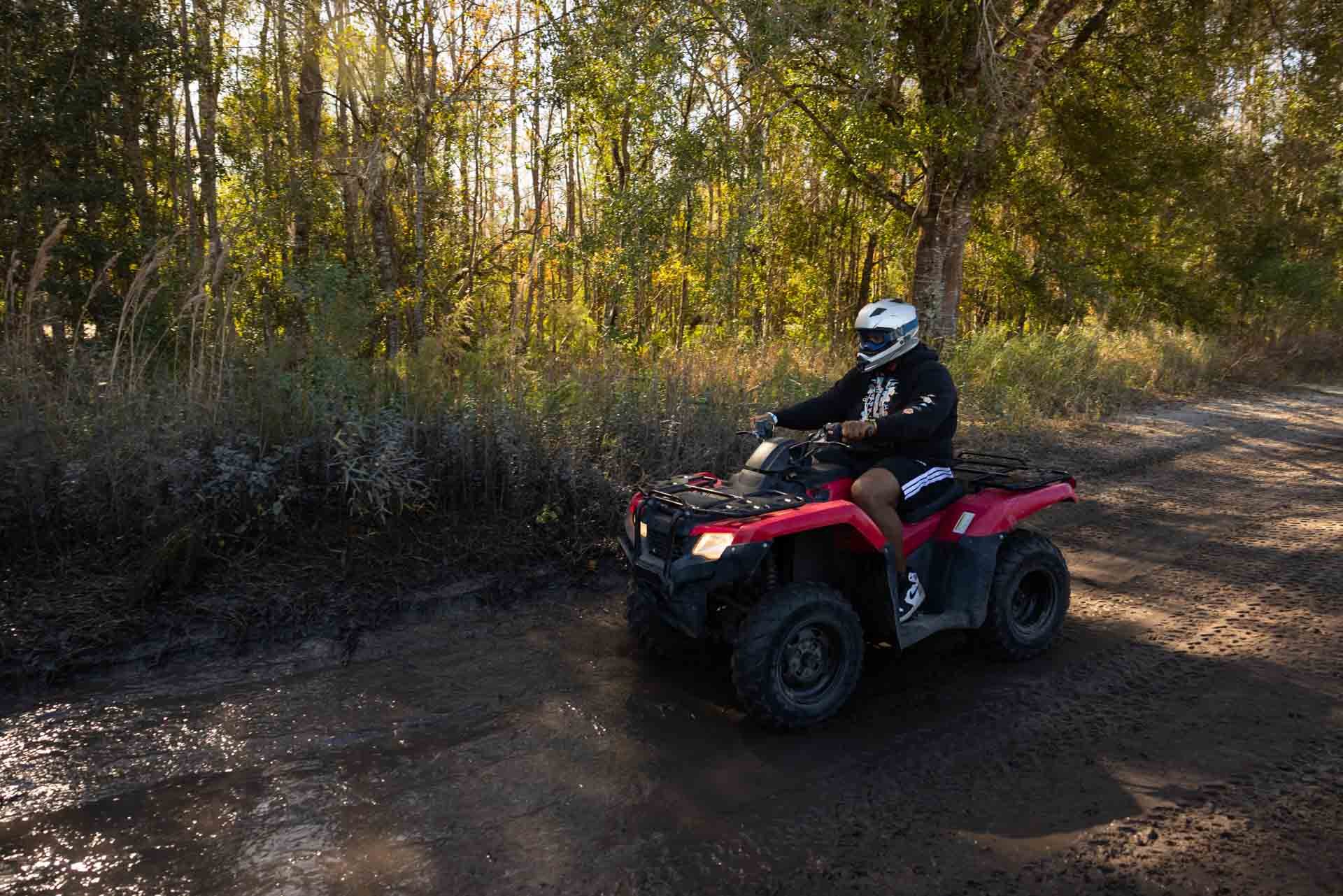 Person riding a red ATV on a dirt trail in a wooded area with sunlight filtering through trees.