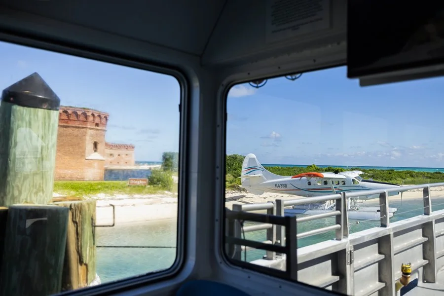 View from inside a boat showing a seaplane docked at a pier, with a historic brick fortress and a sunny shoreline in the background.