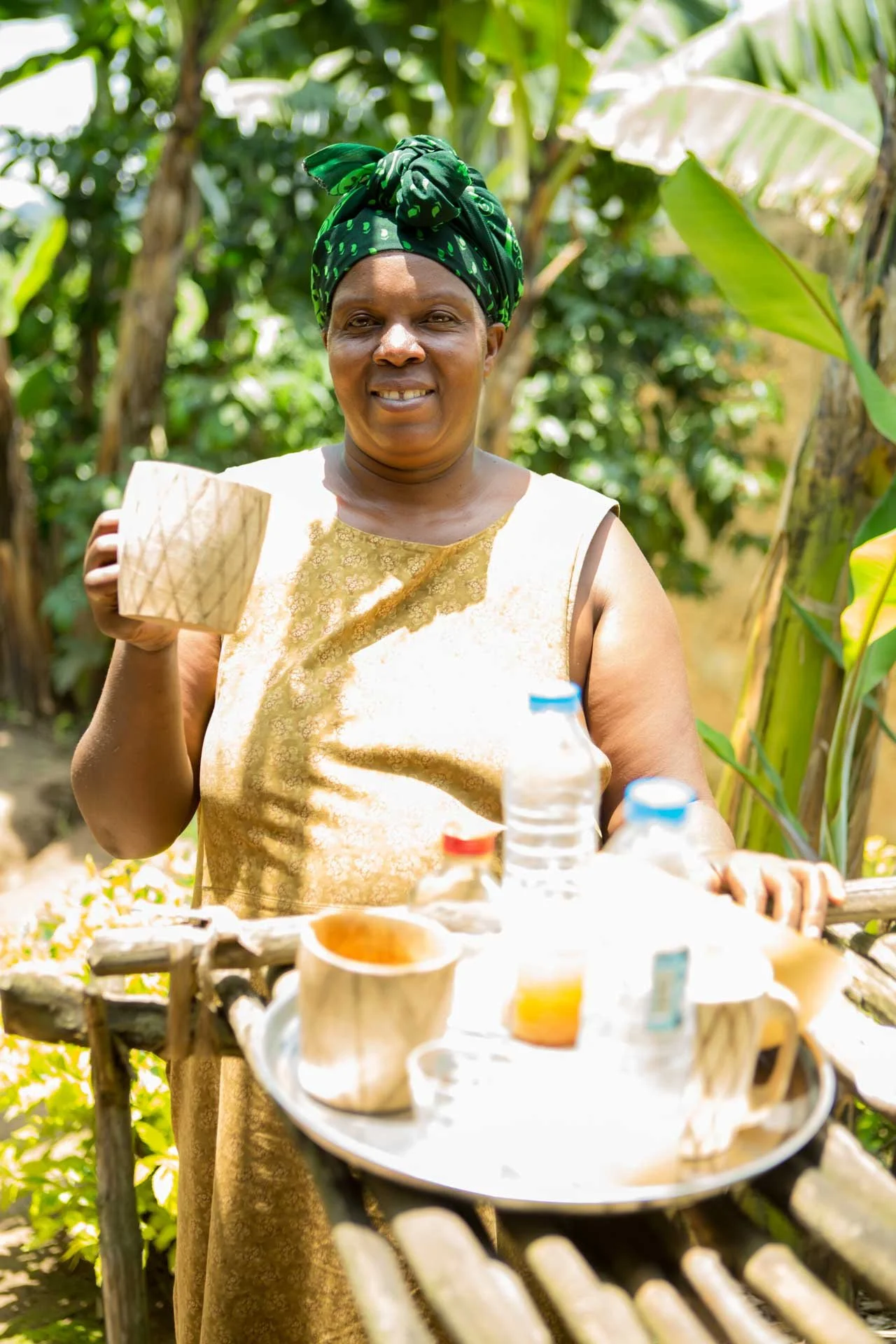 Woman standing outdoors holding a mug, with tea cups and bottles on a tray in front of her, surrounded by green trees.