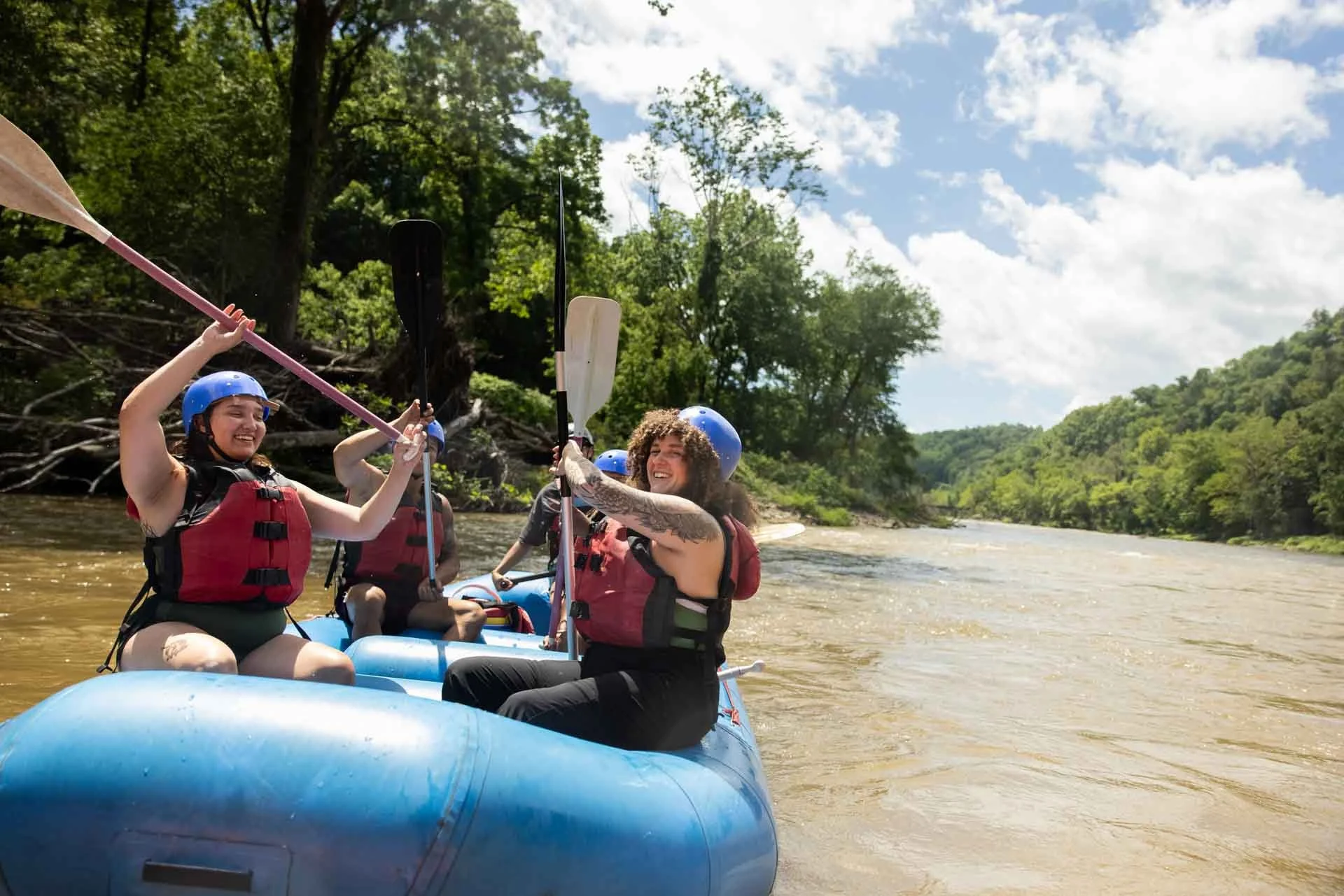 People enjoying white water rafting on a river surrounded by lush green trees on a sunny day.