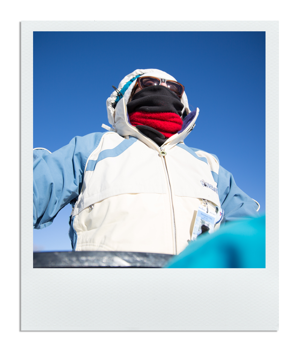 Person wearing winter clothing, sunglasses, and a face covering, outdoors against a clear blue sky.