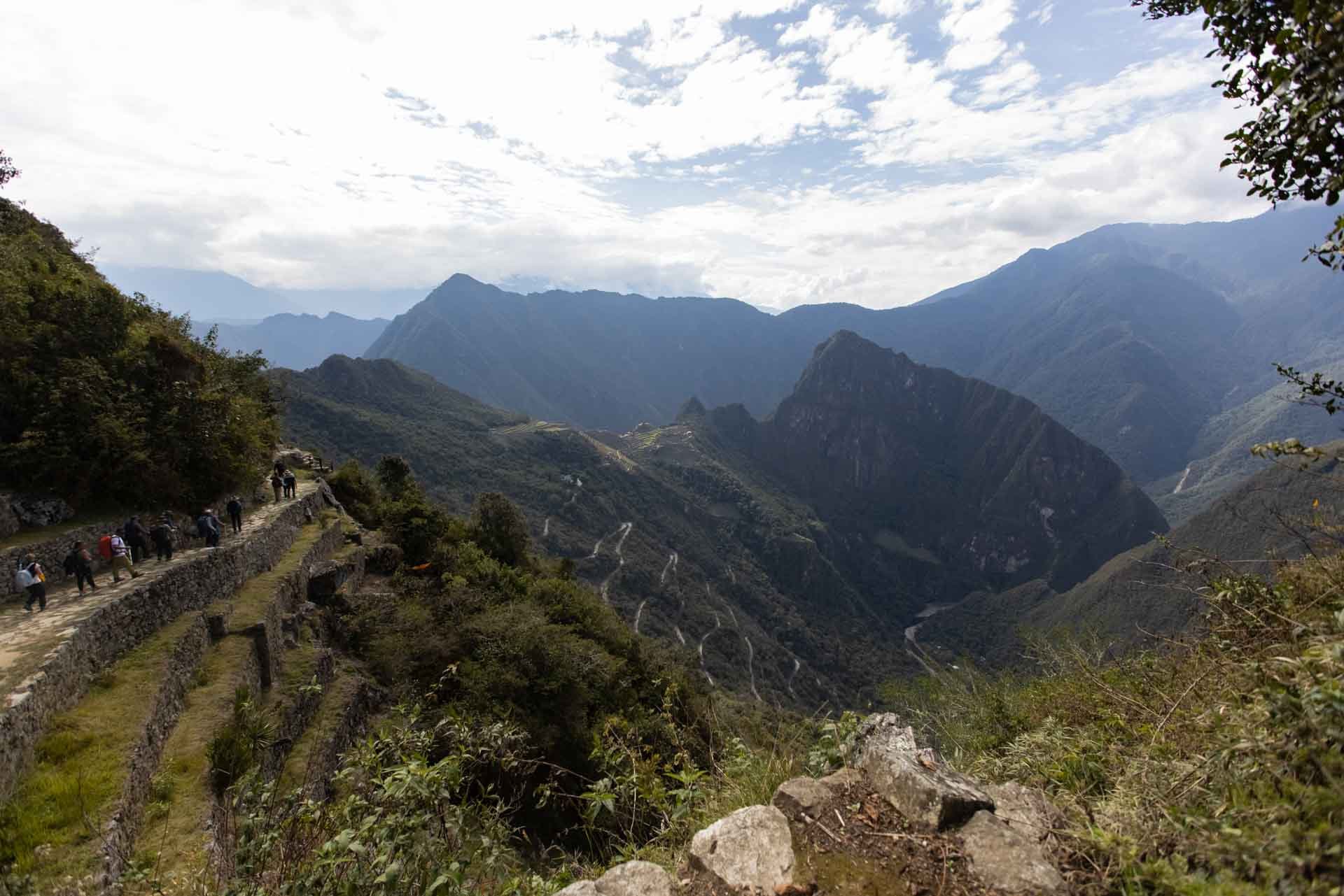 People walking along a mountain trail with lush green hills and towering mountains in the background, partly cloudy sky above.