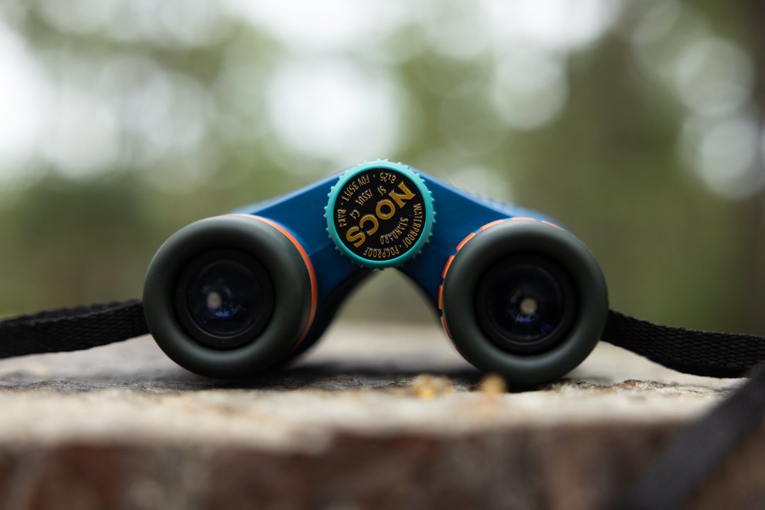 Close-up of colorful binoculars with black lenses, placed on a textured surface outdoors, with a blurred natural background.