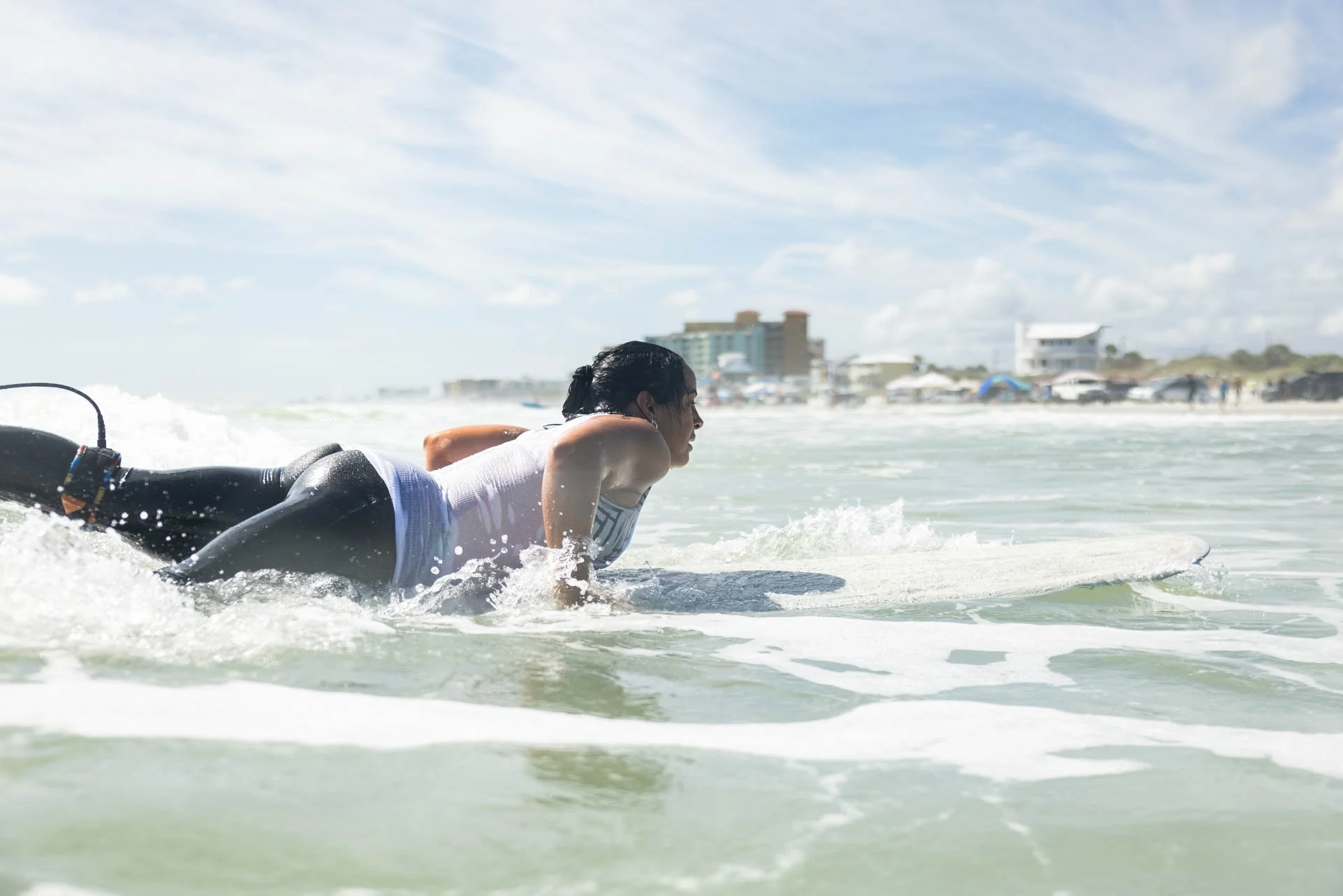 Person lying on a surfboard in the shallow ocean water, preparing to surf near the beach with buildings and umbrellas in the background under a partly cloudy sky.