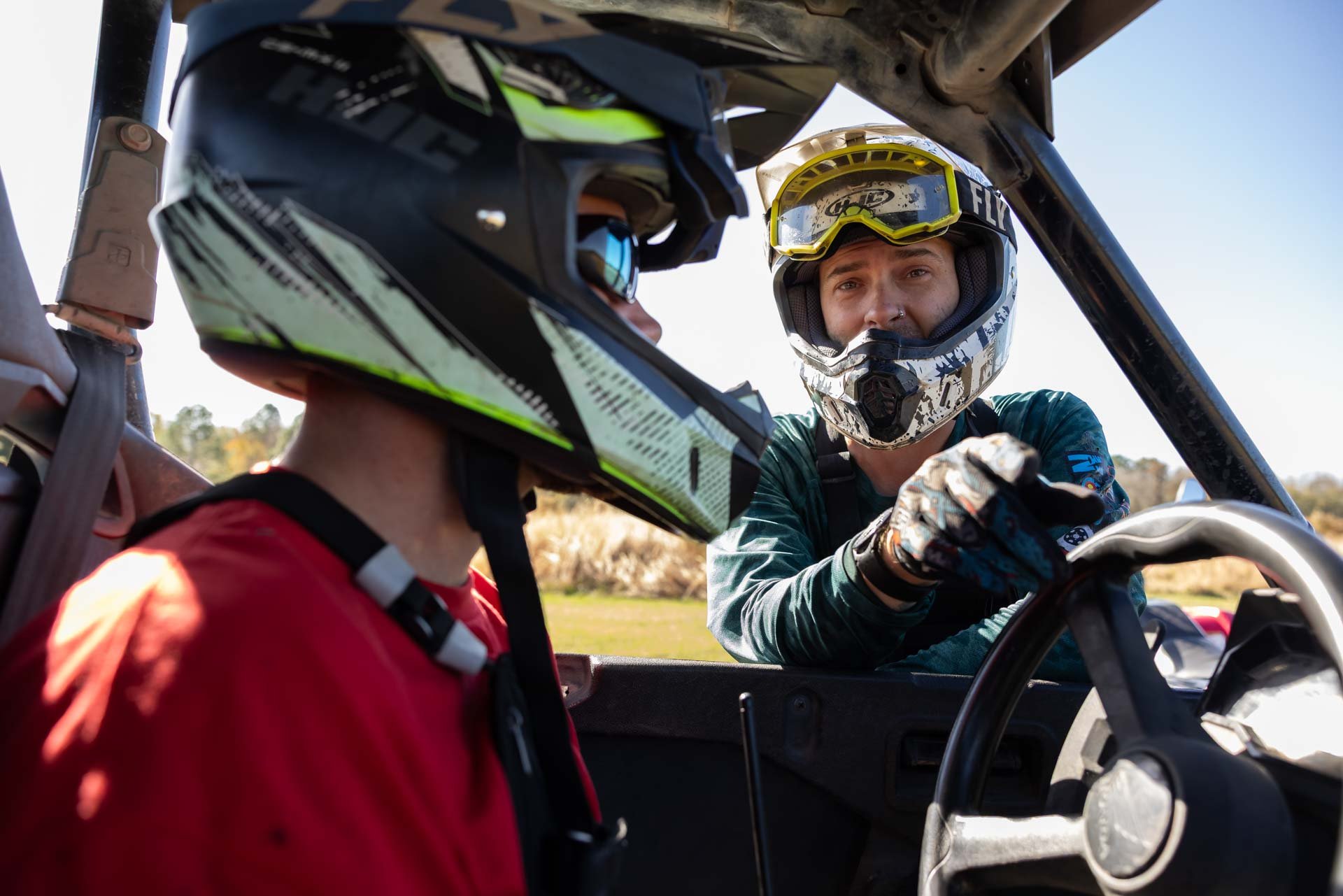 A man in a green racing suit and helmet is talking to a person in a red shirt and black helmet inside a vehicle, with trees and a clear sky in the background.