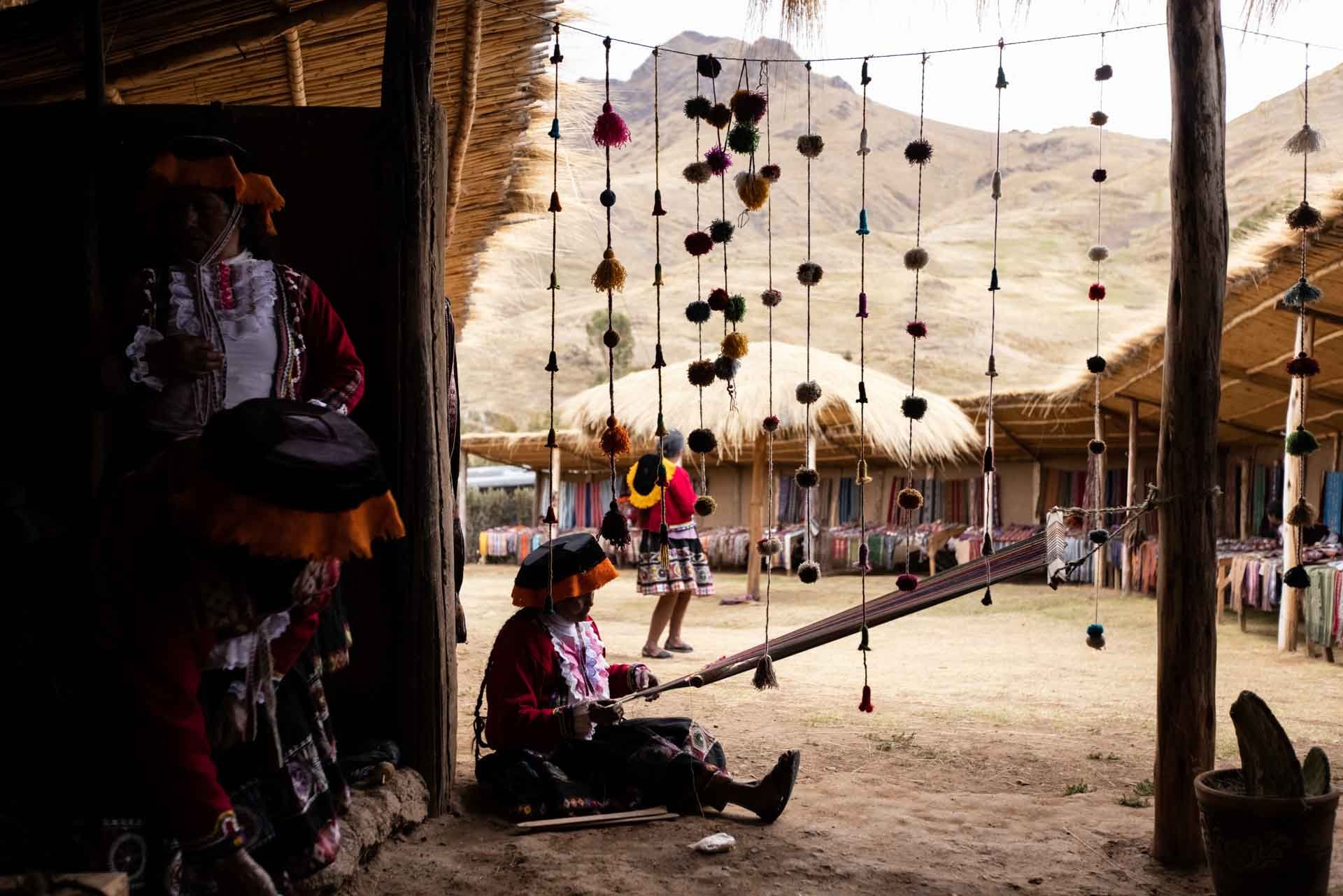 Women dressed in traditional clothing inside a rustic open-air market with hanging decorations and woven tents, mountains visible in the background.
