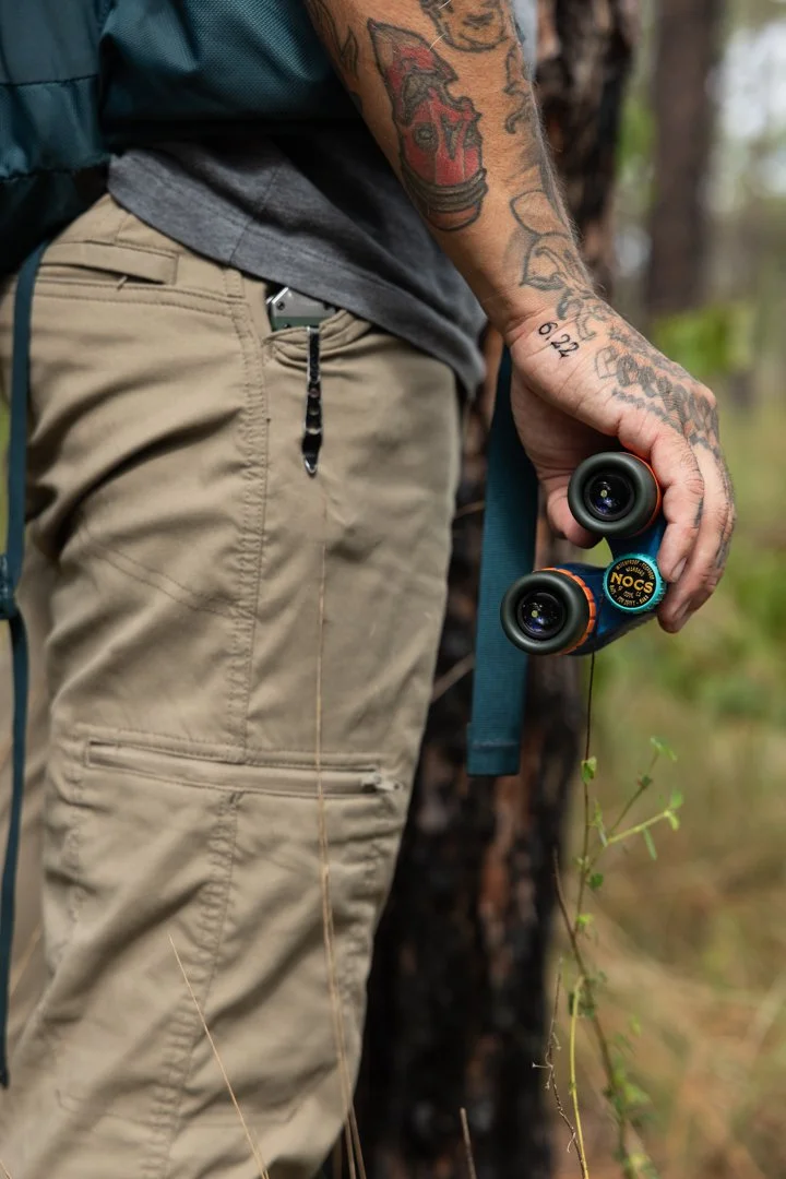 Close-up of a person's hand holding binoculars, with tattoos visible on the arm, standing outdoors in a natural setting with trees and grass.