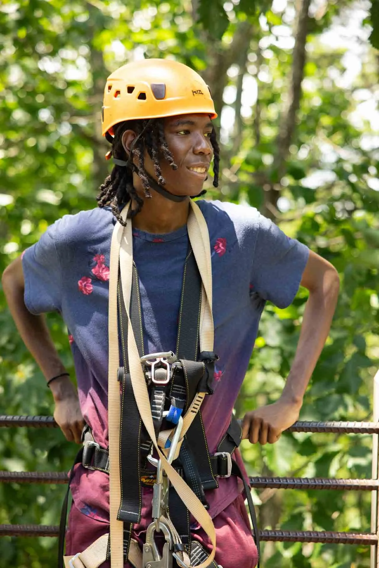 A young man wearing a yellow safety helmet and a harness, standing outdoors among green trees, with a slight smile and a relaxed posture.