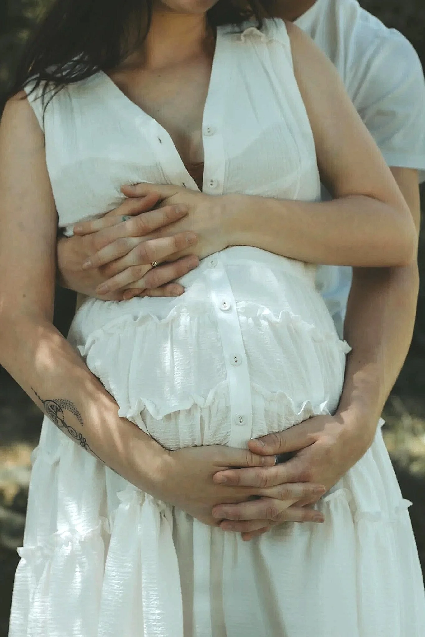 A pregnant woman in a white dress with a person standing behind her, both with their hands cradling her belly.