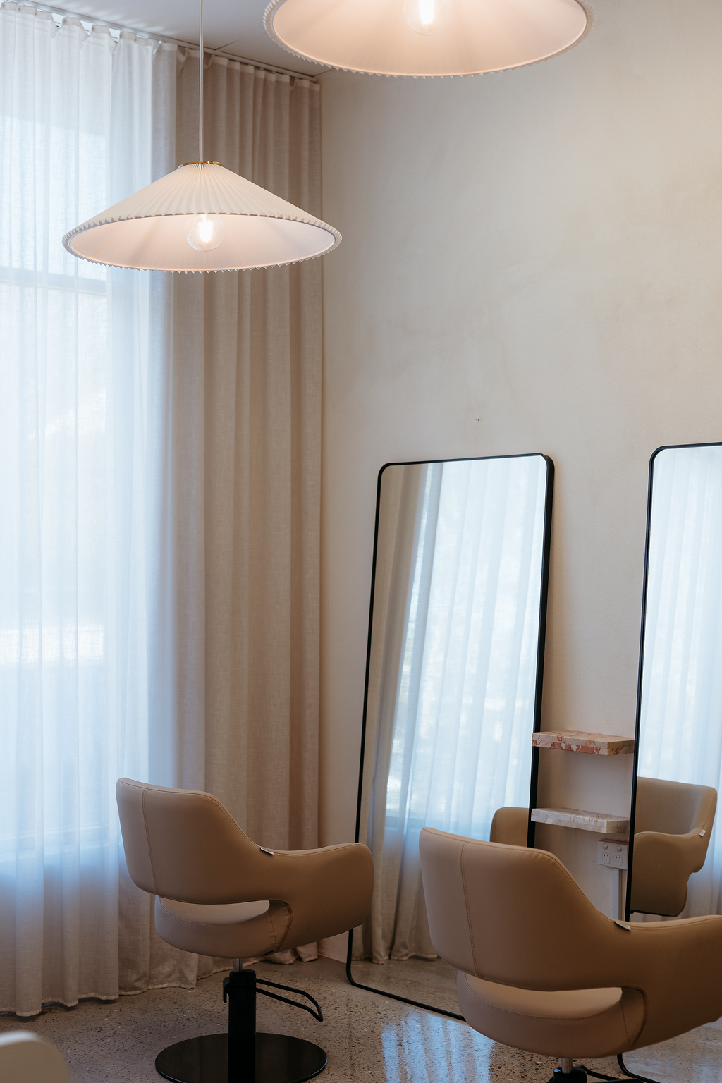 A cozy interior room with two beige salon chairs, two tall freestanding mirrors, beige curtains, and white ceiling lights with shades.