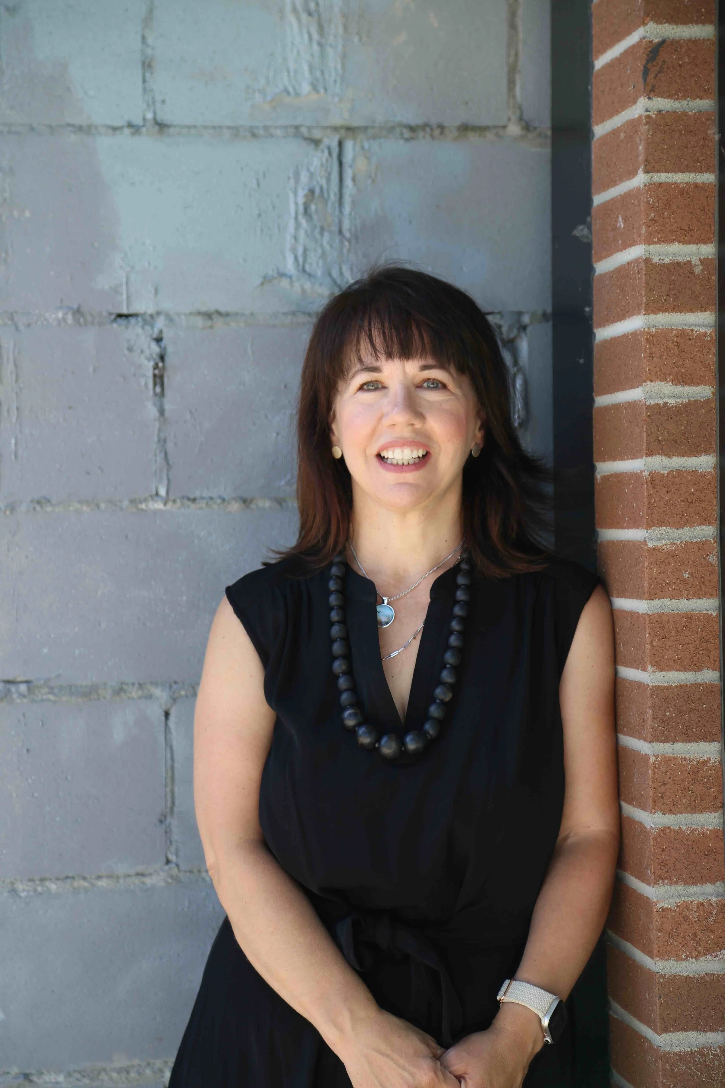 A woman with dark hair, wearing a black dress and necklace, standing outdoors against a concrete and brick wall, smiling at the camera.