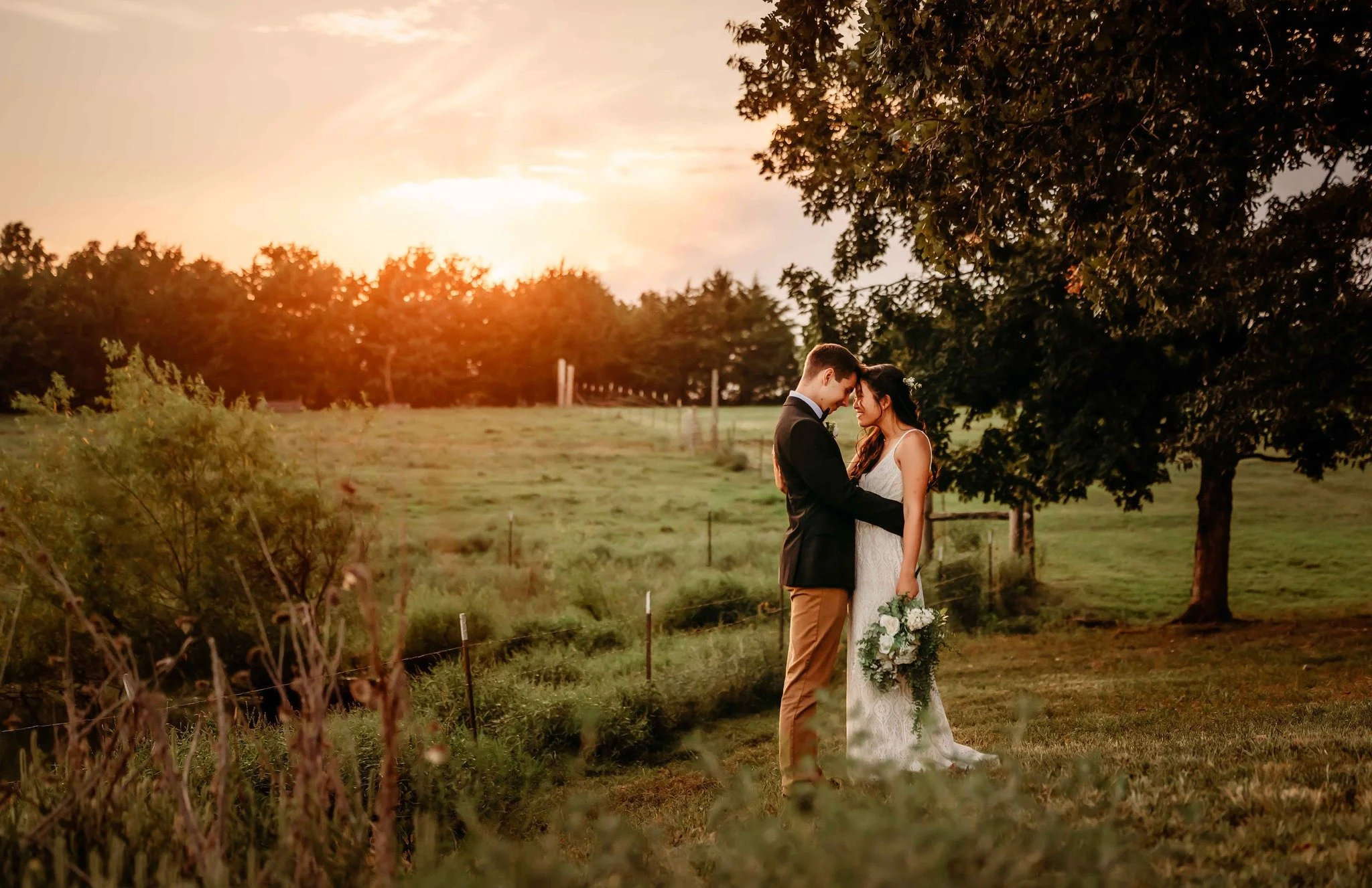 A bride and groom embrace in a grassy outdoor setting during sunset, with trees and a fence in the background.