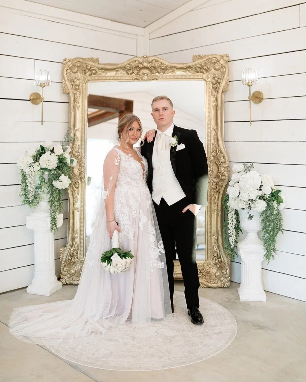 A wedding couple is posing in front of a very large ornate gold floor standing mirror.  The groom is in a black tux and the bride is in her wedding dress with lace and sheer patterns and her flowers are hanging down in her hand.  Serious looks