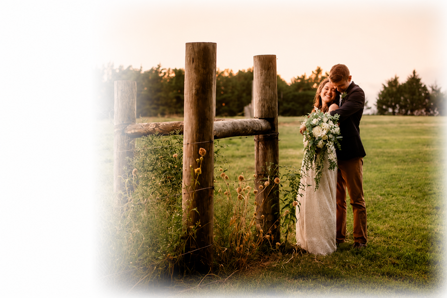 Wedding couple by 3 big wooden post with the groom behind the bride hugging her.  She is in a lace white dress with a huge bouquet of white flowers.