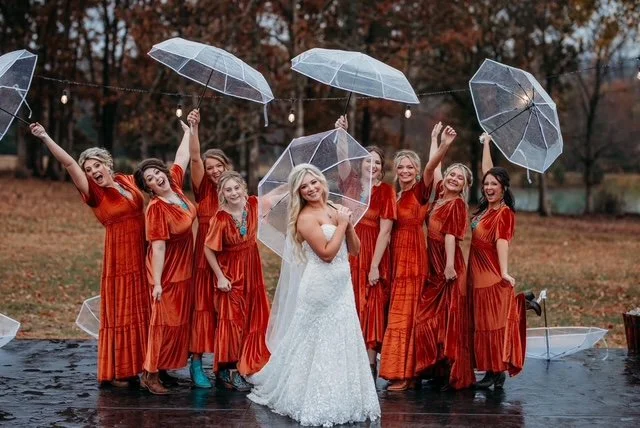 bride at st louis venue in the rain with bridesmaids with umbrellas.  It is an adorable photo with 8 bridesmaids with 4 shared clear umbrellas - the umbrellas are raised in the air and the bride holding her own with two hands leaned on her shoulder