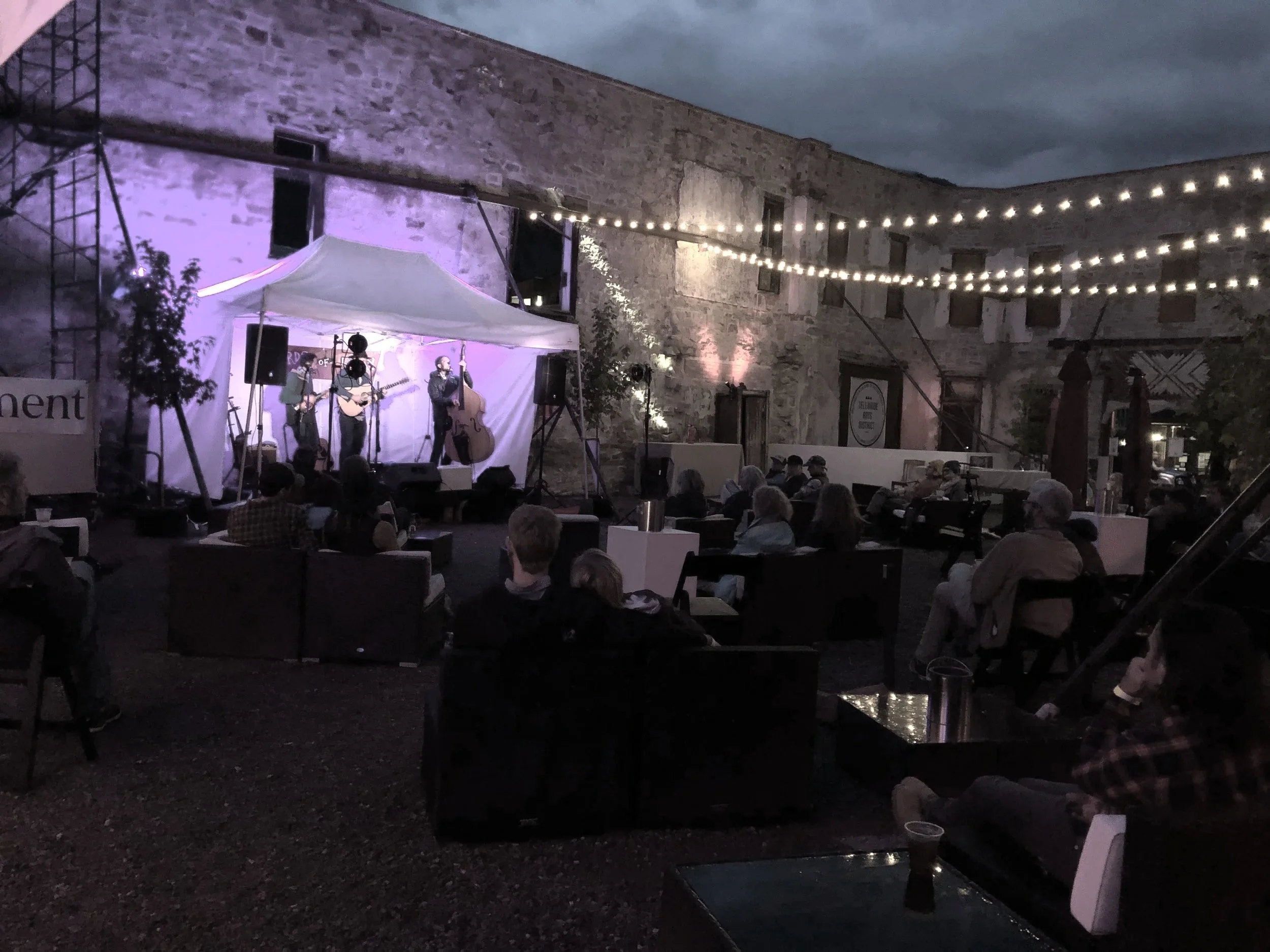 An outdoor evening concert with a small band playing on a stage under a white canopy, audience seated on chairs and couches, lit by string lights against a rustic brick wall.
