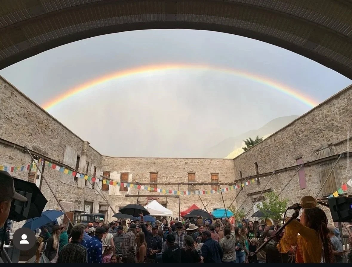 A rainbow over an outdoor gathering in a courtyard with stone walls, decorated with colorful banners, people holding umbrellas, and a singer performing at a microphone.